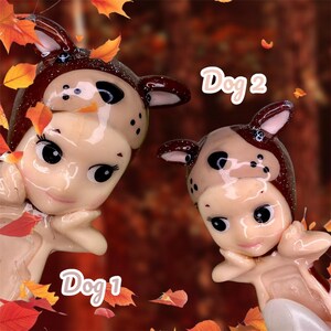 Pu&ograve; includere: Due piccole bambole di porcellana vestite da cani. La bambola a sinistra &egrave; etichettata "Dog 1" e la bambola a destra &egrave; etichettata "Dog 2". Entrambe le bambole indossano costumi da cane marroni e bianchi con accenti di glitter. Le bambole sono circondate da foglie autunnali.