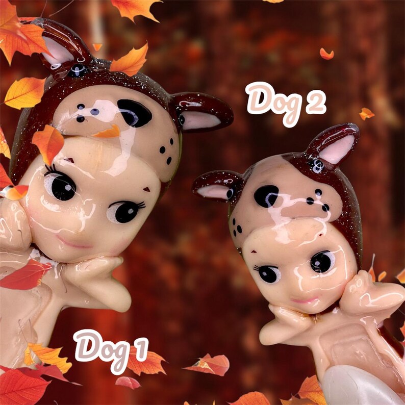 Pu&ograve; includere: Due piccole bambole di porcellana vestite da cani. La bambola a sinistra &egrave; etichettata "Dog 1" e la bambola a destra &egrave; etichettata "Dog 2". Entrambe le bambole indossano costumi da cane marroni e bianchi con accenti di glitter. Le bambole sono circondate da foglie autunnali.