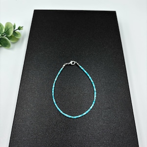 May include: A delicate turquoise bead bracelet with a silver clasp. The bracelet is made of small, evenly sized, light blue beads. The clasp is a small, silver-coloured lobster claw style. The bracelet is displayed on a black surface.