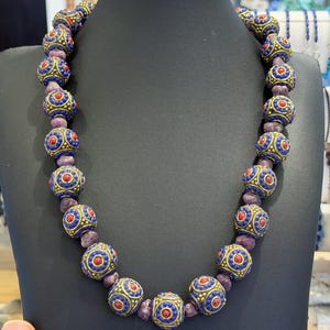 May include: A necklace featuring alternating round beads. The larger beads are crafted from brass with intricate blue and red enamel detailing, while the smaller, irregular spacer beads are a deep, textured purple stone.