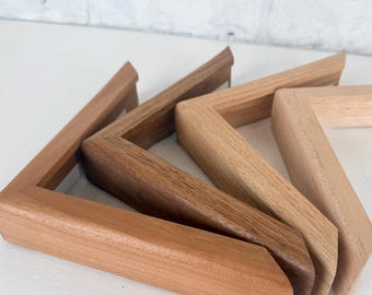May include: Four L-shaped wooden picture frame corner samples arranged in a row. Each piece displays a different wood grain and natural tone, ranging from a light pale wood to a medium brown and a darker, rich walnut-colored wood finish.