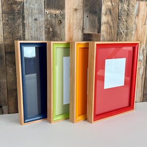 May include: Four colorful picture frames with natural wood-colored borders. The frames are navy blue, green, orange, and red. Each frame has a white square in the center. The frames are leaning against a wooden wall.