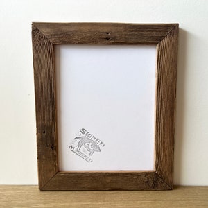 May include: A rustic, rectangular picture frame made of weathered, dark brown wood. The frame has a white interior and a stamp in the lower right corner that reads "SIGNED NUMBERED". The frame is sitting on a light brown surface.