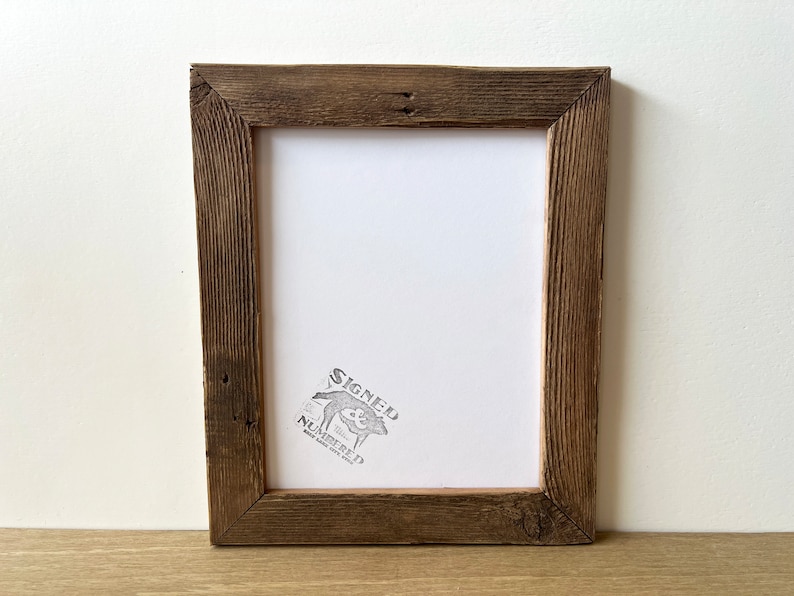 May include: A rustic, rectangular picture frame made of weathered, dark brown wood. The frame has a white interior and a stamp in the lower right corner that reads "SIGNED NUMBERED". The frame is sitting on a light brown surface.