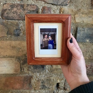 May include: A small, rectangular wooden picture frame with a brown finish. The frame holds a polaroid photo of two people. The frame is held in front of a brick wall.