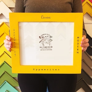 May include: A yellow wood picture frame with the words "Cursive", "Handwriting", "Modern", and "Typewriter" written on it. The frame has a white mat and a black and white stamp that says "Handmade in USA Signed Numbered Salt Lake City 100% Solid Hardwood".