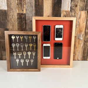 May include: Two framed shadow boxes. One contains an assortment of vintage keys in various shapes and sizes. The other holds four smartphones, two with cracked screens, against a red background. Both frames have wooden borders.