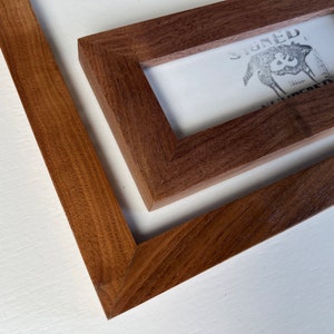 May include: Close-up of two wooden picture frames. The frames are a rich brown color and have a simple, modern design. One frame contains a print with the text "SIGNED & NUMBERED" and a graphic. The frames are set against a white background.