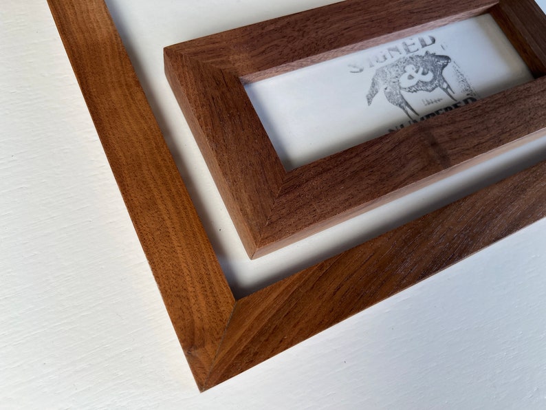 May include: Close-up of two wooden picture frames. The frames are a rich brown color and have a simple, modern design. One frame contains a print with the text "SIGNED & NUMBERED" and a graphic. The frames are set against a white background.
