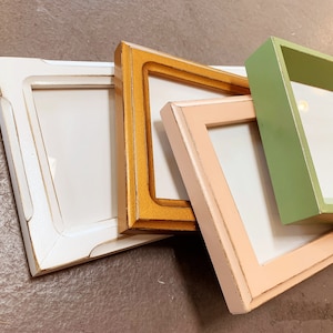 May include: Four different picture frames in various colors, including white, light pink, light green, and gold. The frames are stacked on top of each other, with the white frame on the bottom and the gold frame on top.