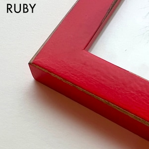 May include: Close-up of the corner of a red wooden picture frame. The frame has a distressed finish and a slightly raised edge.