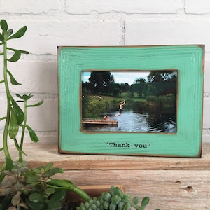 May include: A teal wooden picture frame with a photo of a person jumping into a lake. The frame has the text "Thank you" written on the bottom.