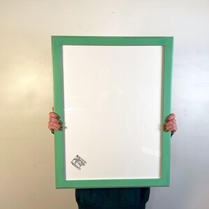 May include: A rectangular picture frame with a smooth, light green finish. The frame surrounds a large, blank white mat board. In the bottom left corner of the white mat, there is a small, black ink stamp featuring a drawing of a pig.