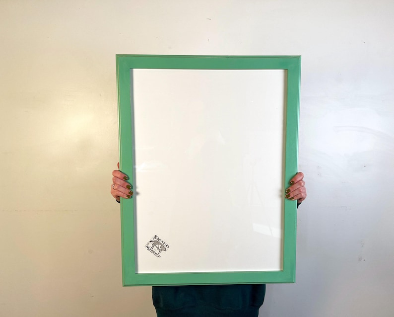 May include: A rectangular picture frame with a smooth, light green finish. The frame surrounds a large, blank white mat board. In the bottom left corner of the white mat, there is a small, black ink stamp featuring a drawing of a pig.