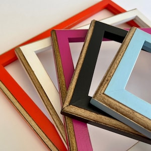 May include: Five different colored picture frames with a wood grain finish. The frames are stacked on top of each other, with the largest frame on the bottom and the smallest frame on top. The frames are painted in shades of orange, white, pink, black, and light blue.