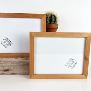 May include: Two rectangular light brown wooden picture frames with white inserts. Each insert features a small black graphic of a fox with the text SIGNED NUMBERED printed in a circular pattern. The frames are positioned at different heights.