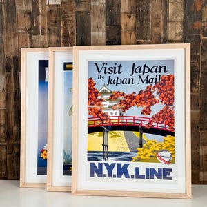 May include: Three framed travel posters in light wood frames. The front poster features a red bridge, a white castle, and autumn foliage with the text Visit Japan By Japan Mail and N.Y.K.LINE. The posters are displayed in a row on a white surface.