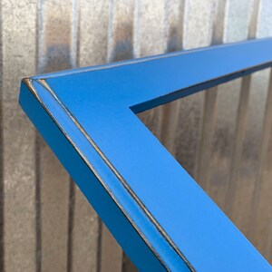 May include: A bright blue rectangular picture frame with a smooth finish. The edges of the frame feature a distressed look, revealing dark brown wood tones along the corners and outer borders, highlighting the construction of the frame.