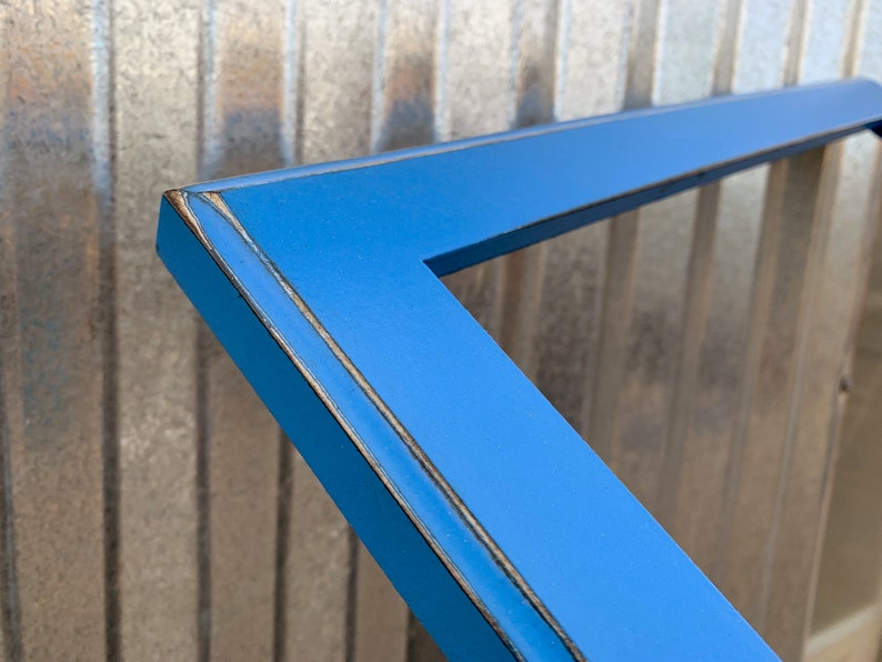 May include: A bright blue rectangular picture frame with a smooth finish. The edges of the frame feature a distressed look, revealing dark brown wood tones along the corners and outer borders, highlighting the construction of the frame.