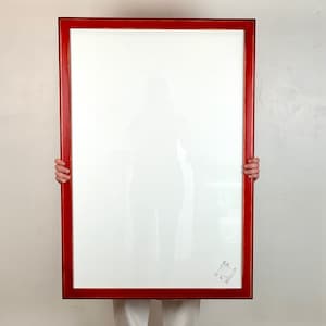 May include: A large rectangular picture frame with a glossy red finish and a white mat board. The frame features a clear glass front and a small, square-shaped decorative emblem or logo printed in the bottom right corner of the white matting.