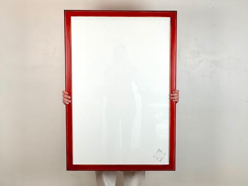 May include: A large rectangular picture frame with a glossy red finish and a white mat board. The frame features a clear glass front and a small, square-shaped decorative emblem or logo printed in the bottom right corner of the white matting.