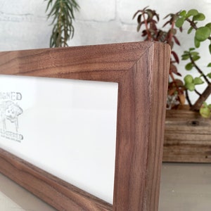 May include: A rectangular wooden picture frame with a light-colored interior and a dark brown, textured exterior. The frame has the words "SIGNED" and "NUMBERED" printed on the interior. The frame is displayed on a white surface.