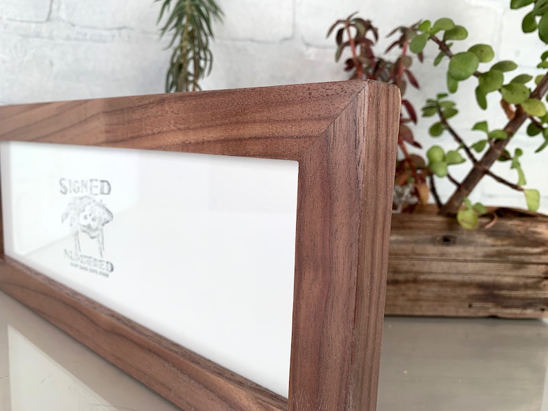 May include: A rectangular wooden picture frame with a light-colored interior and a dark brown, textured exterior. The frame has the words "SIGNED" and "NUMBERED" printed on the interior. The frame is displayed on a white surface.