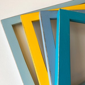 May include: Four wooden picture frames in different colors: light blue, yellow, blue, and teal. The frames are stacked on top of each other, with the light blue frame on the bottom and the teal frame on top.