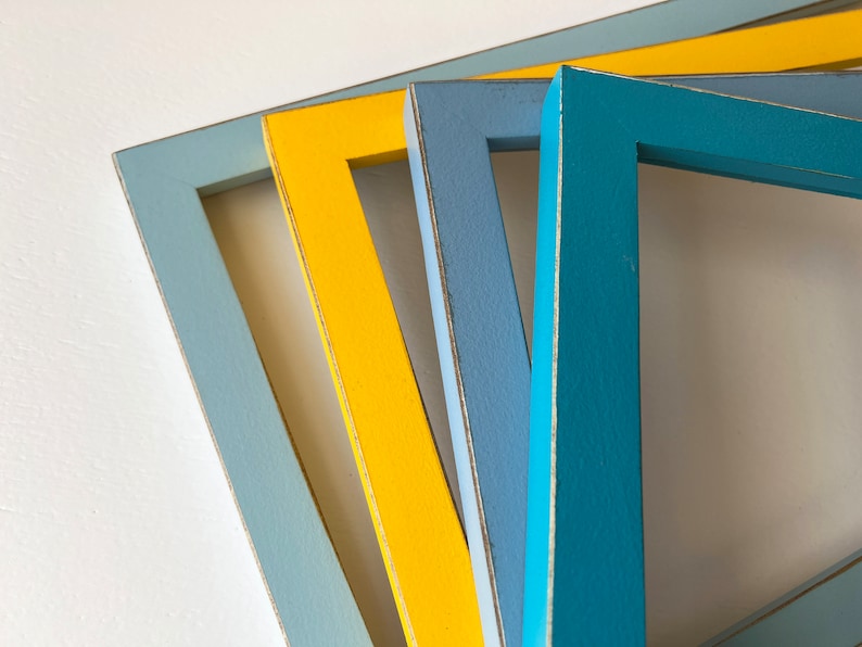 May include: Four wooden picture frames in different colors: light blue, yellow, blue, and teal. The frames are stacked on top of each other, with the light blue frame on the bottom and the teal frame on top.