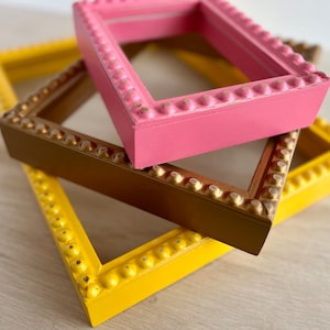 May include: Three picture frames stacked on top of each other. The top frame is pink, the middle frame is gold, and the bottom frame is yellow. All three frames have a beaded design around the edges.