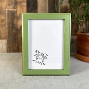 May include: A rectangular picture frame with a light green finish and distressed edges. The frame contains a white insert featuring a black graphic of a wolf and the text "SIGNED & NUMBERED" along with smaller text below the graphic.