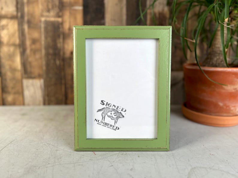May include: A rectangular picture frame with a light green finish and distressed edges. The frame contains a white insert featuring a black graphic of a wolf and the text "SIGNED & NUMBERED" along with smaller text below the graphic.