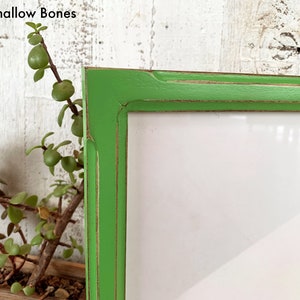 May include: A bright green picture frame with a distressed finish showing brown wood underneath the paint. The frame has a rectangular profile and contains a blank white insert. The text 1x1 Shallow Bones is visible in the upper left corner.