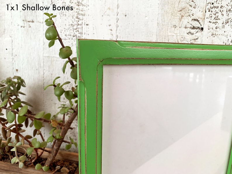 May include: A bright green picture frame with a distressed finish showing brown wood underneath the paint. The frame has a rectangular profile and contains a blank white insert. The text 1x1 Shallow Bones is visible in the upper left corner.