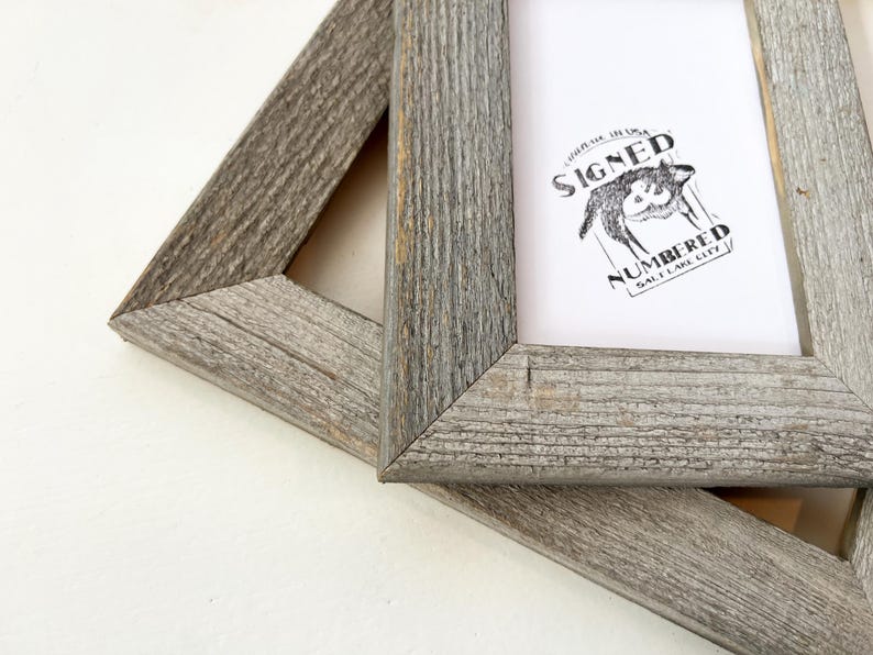 May include: Two weathered gray wooden picture frames. One frame contains a white print with a black illustration of a wolf and the words "SIGNED" and "NUMBERED" above and below the wolf, respectively. The frames are angled.