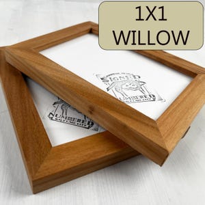 May include: Two wooden picture frames, one on top of the other, both with a light brown finish. The top frame is slightly larger than the bottom frame. The frames are empty, but the top frame has a white paper insert with the text "KIPMAN IN USA SIGNED NUMBERED SALT LAKE CITY" printed on it.