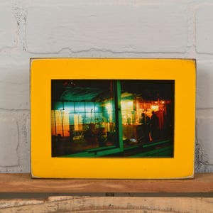 May include: A yellow picture frame with a photograph of a brightly lit storefront at night. The storefront has a green and blue awning and a red and yellow sign.