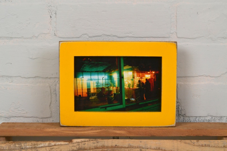 May include: A yellow picture frame with a photograph of a brightly lit storefront at night. The storefront has a green and blue awning and a red and yellow sign.