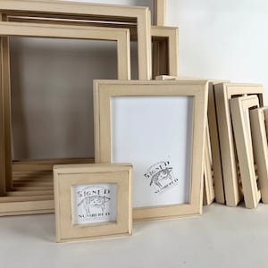 May include: A collection of beige wooden picture frames of various sizes, stacked and arranged on a white surface. The frames are all made of the same material and have a simple, classic design. The largest frame is in the back, and the smallest frame is in the front. The frames are all empty, and there is no artwork or photos inside them.
