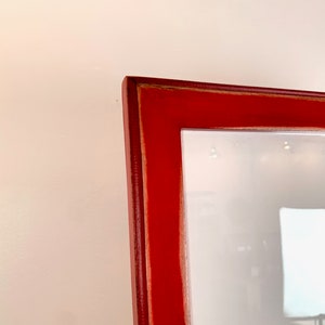 May include: A close-up view of the corner of a rectangular picture frame with a smooth, vibrant red finish. The frame features a mitered joint at the corner, showing a thin, light-colored wood edge along the inner seam of the red painted surface.