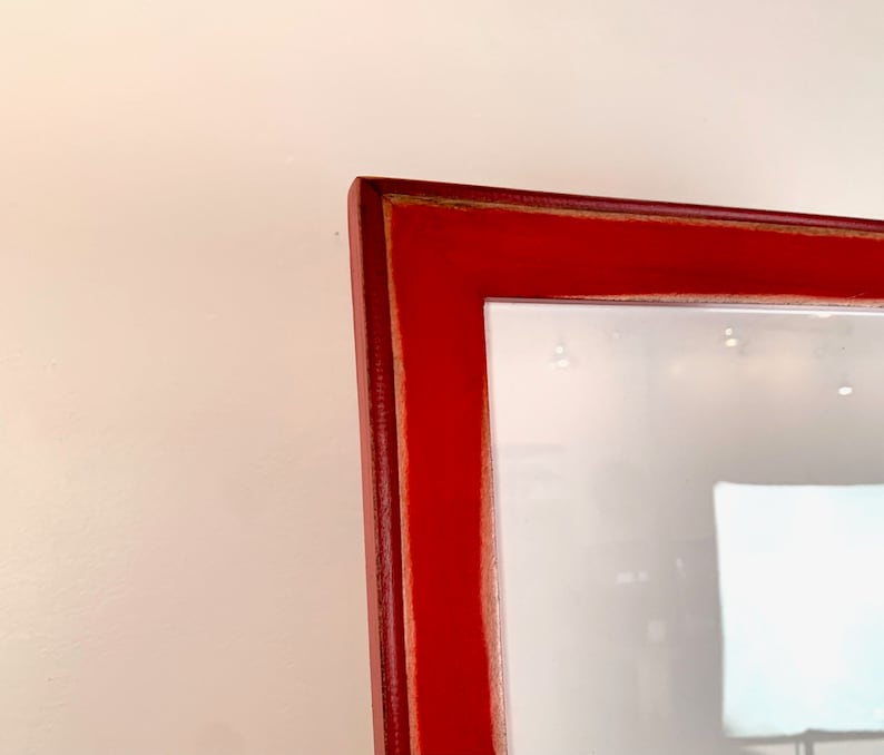 May include: A close-up view of the corner of a rectangular picture frame with a smooth, vibrant red finish. The frame features a mitered joint at the corner, showing a thin, light-colored wood edge along the inner seam of the red painted surface.