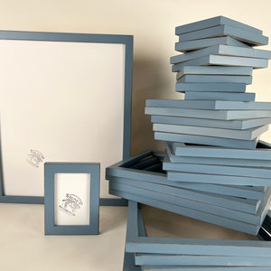 May include: A stack of blue picture frames, some are stacked vertically and some are stacked horizontally. There is a larger blue picture frame to the left of the stack and a smaller blue picture frame in front of the stack. The frames are all made of wood and have a distressed finish.