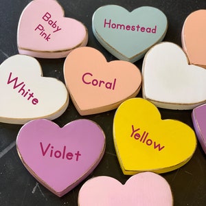 May include: A collection of wooden heart-shaped ornaments painted in various colors, including baby pink, coral, white, homestead blue, yellow, violet, and pink. Each heart has a gold trim and a different color name written on it.