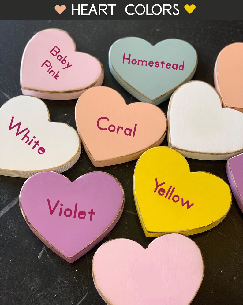 May include: A collection of wooden heart-shaped ornaments painted in various colors, including baby pink, coral, white, homestead blue, yellow, violet, and pink. Each heart has a gold trim and a different color name written on it.