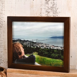 May include: A framed photograph of a young person looking out at a view of a city and a bridge over a body of water. The photo is in a brown frame.