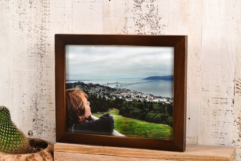 May include: A framed photograph of a young person looking out at a view of a city and a bridge over a body of water. The photo is in a brown frame.