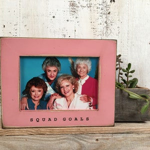 May include: Pink wooden picture frame with a photo of four women inside. The frame has the words "SQUAD GOALS" printed on the bottom.