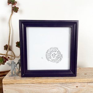 May include: A square, dark purple picture frame with a white interior. The frame has a simple, modern design. The text "SIGNED & NUMBERED SALT LAKE CITY" is printed in a circle inside the frame. The frame is displayed on a wooden surface.