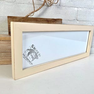 May include: A rectangular, cream-colored wooden picture frame with a clear glass front. Inside the frame, a white paper insert features a black graphic illustration of a bison with the text "SIGNED & NUMBERED" printed in a bold, stylized font.