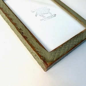 May include: A rectangular picture frame with a weathered, sage green finish and visible wood grain texture. The frame contains a white insert featuring the text SIGNED NUMBERED in black ink, accompanied by a small graphic of a pig illustration.